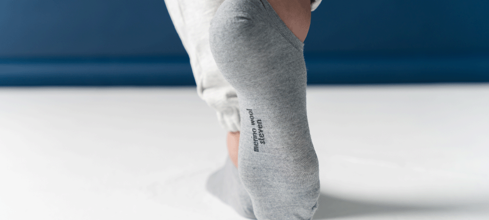 Close-up of a person wearing grey merino wool STEVEN no-show socks, with the brand name visible on the sole, against a white and blue background